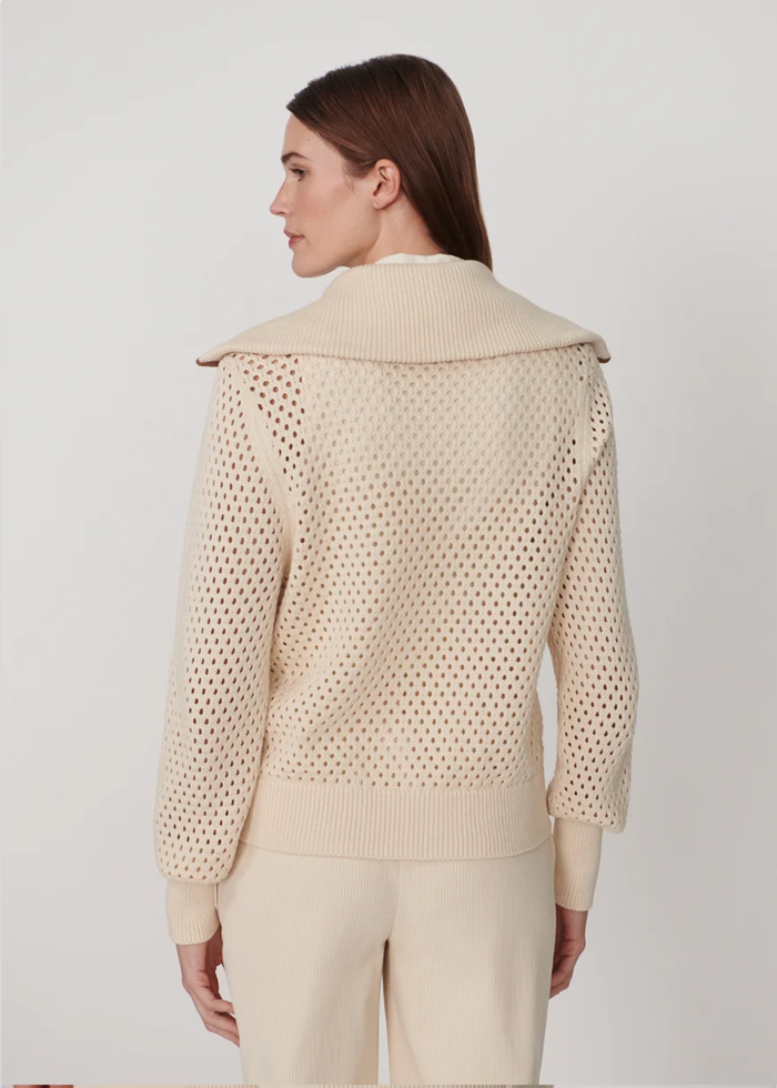 A woman shown from the back wearing a cream-colored knit sweater with a wide collar and openwork knit pattern, paired with matching cream pants, against a plain light gray studio background.