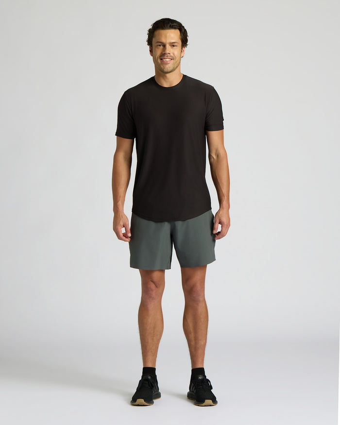 Man wearing an All Black Active Curved Hem Crew 6-Pack black short sleeve crew neck t-shirt paired with gray athletic shorts and black sneakers, standing against a plain background.
