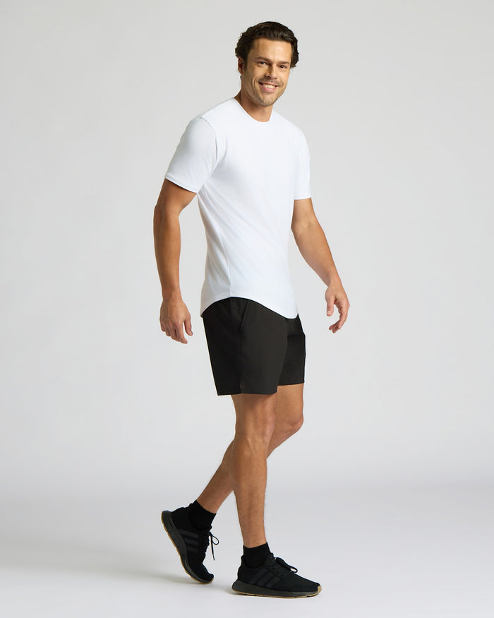 Man wearing All White Active Curved Hem Crew 6-Pack t-shirt, paired with black shorts and sneakers, smiling while walking against a plain light background.