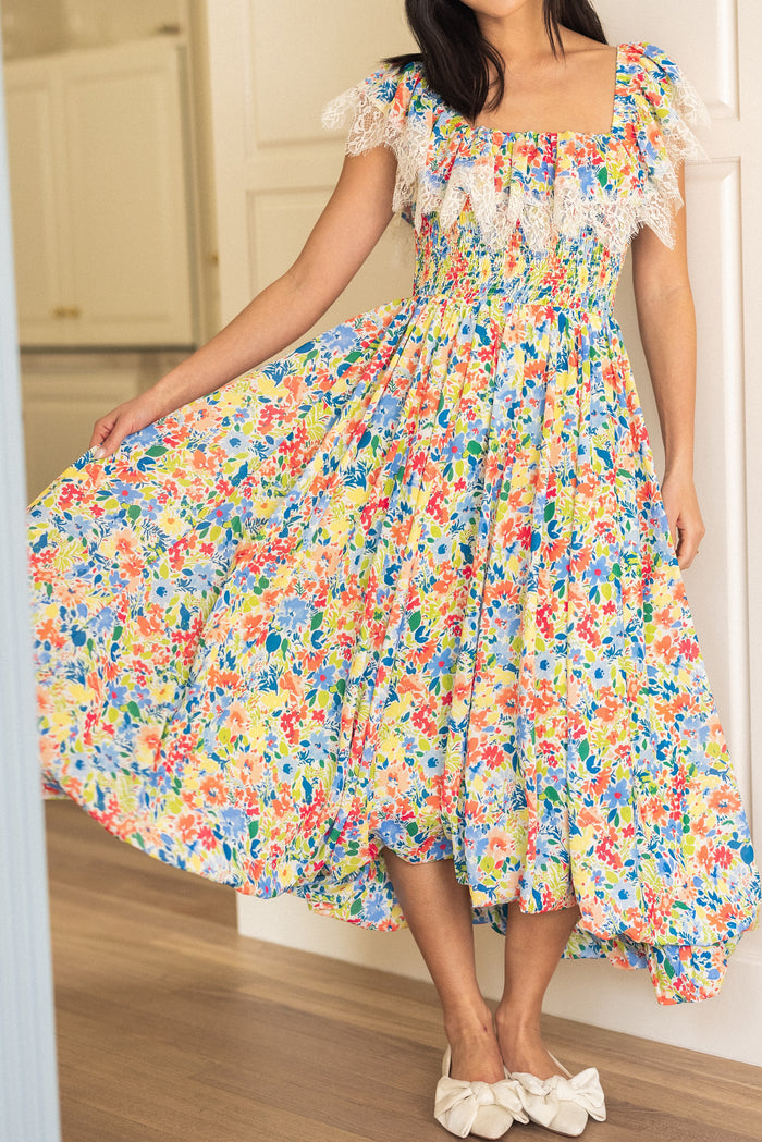 A woman with long dark hair wears the Ivy City Co Ada Dress in Vibrant Yarrow Print, an ankle-length, nursing-friendly chiffon dress with lace trim, styled with white bow shoes as she stands indoors holding out her skirt.