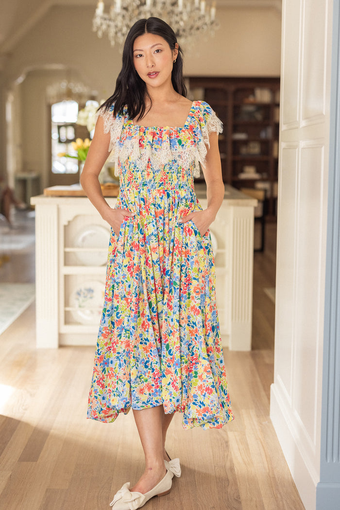 A woman in Ivy City Co's Ada Dress in Vibrant Yarrow Print—a colorful floral chiffon midi with lace ruffle neckline, smocked bodice, pockets, and maternity-friendly fit—pairs it with white bow flats in an elegant interior.