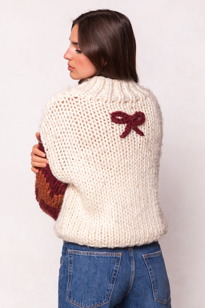 A woman with long brown hair wears Ivy City Co’s Aimee Sweater in Orange & Cream—an oversized wool blend with a small maroon bow on the upper back—paired with blue jeans and turned sideways to showcase the detail.