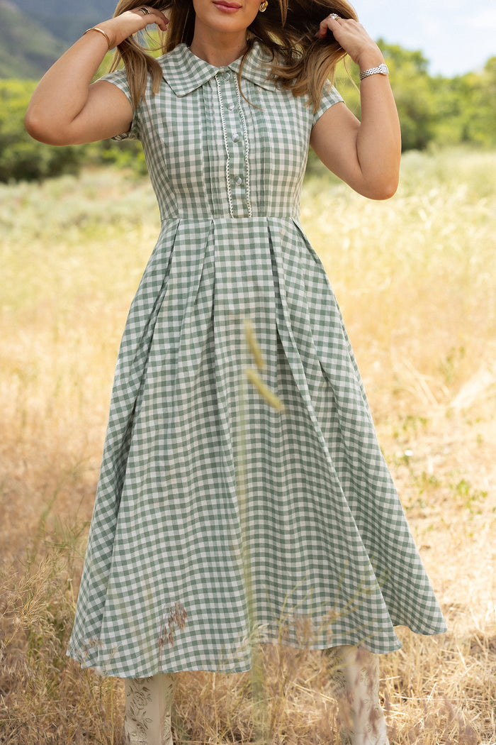 A woman stands in a sunlit field wearing the Ivy City Co Amber Dress in Sage Gingham, featuring short sleeves, a collar, and a pleated cotton midi skirt. Grass and trees fill the background as her face is partially out of frame.