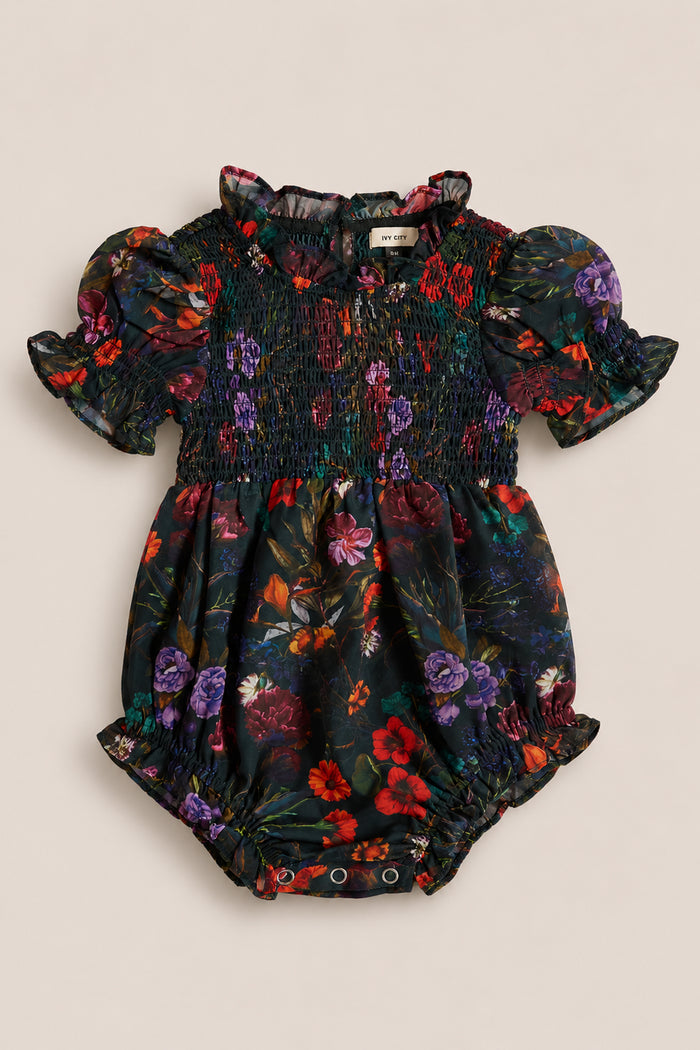 The Ivy City Co Baby Cordelia Romper in Evergreen Vivid Meadow is a charming dark floral baby romper, featuring puffed short sleeves, a smocked bodice, ruffled collar, and convenient snap buttons at the bottom—perfect as a cozy fall baby outfit.