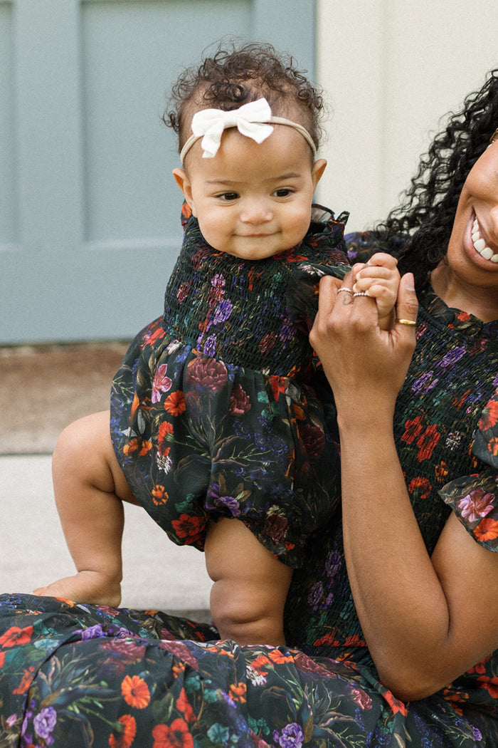 A smiling baby wears the adorable Ivy City Co Baby Cordelia Romper in Evergreen Vivid Meadow—a floral fall baby outfit—paired with a white bow headband, and is held by an adult who is partially visible and also dressed in a matching floral outfit.