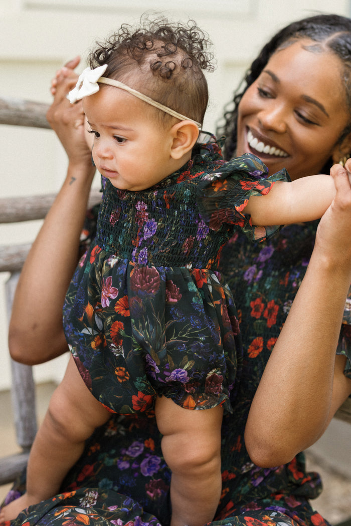 A smiling woman holds up a baby wearing the Ivy City Co Baby Cordelia Romper in Evergreen Vivid Meadow—a charming fall baby outfit featuring a floral dress and a white bow headband. Both are dressed in matching dark floral outfits, sitting together on a wooden bench.