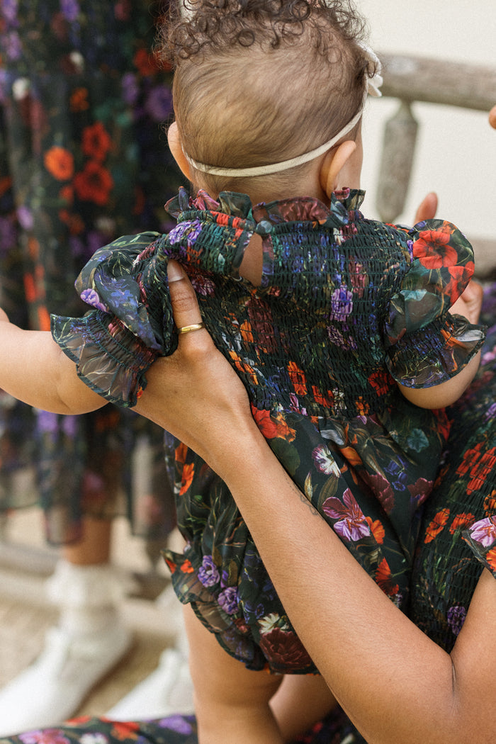 An infant wearing the Ivy City Co Baby Cordelia Romper in Evergreen Vivid Meadow, featuring dark floral fabric and ruffled sleeves, is held by an adult dressed in a coordinating floral dress. The baby's look is completed with a white headband, while the adult's supportive embrace showcases their charming fall outfit.