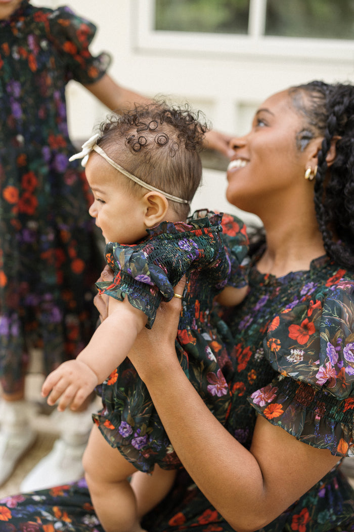 A smiling woman cradles a baby in her arms, the child dressed in Ivy City Co’s Baby Cordelia Romper in Evergreen Vivid Meadow—a charming fall outfit with a floral pattern that complements the woman's own dress. Nearby, another child, also wearing a similar Baby Cordelia Romper, stands partially out of frame.