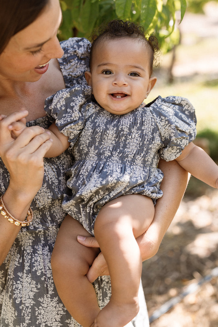 A smiling baby wears the Ivy City Co Baby Lisa Romper in Jolie Fleur, featuring a smocked bodice and puff sleeves, while being held by an adult in a matching dress. They are outdoors with greenery in the background.