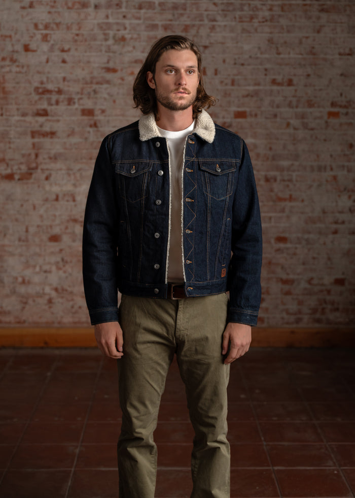 Man in a denim jacket with a shearling collar stands against a brick wall, wearing khaki pants and a white shirt, looking forward.