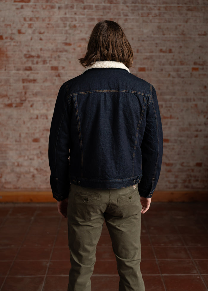 A person with long hair wears a denim jacket with a shearling collar, standing in front of a textured brick wall.
