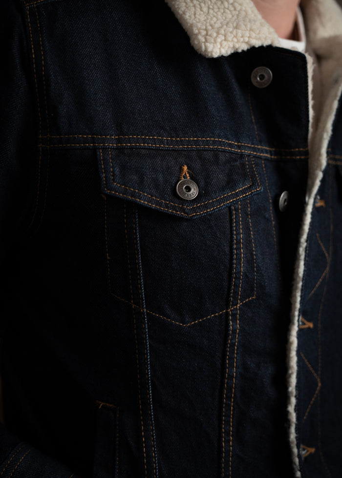 Close-up of a dark denim jacket with a white sherpa collar, highlighting the buttoned pocket and detailed stitching.
