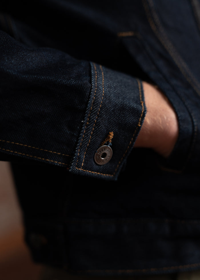 A hand rests in the pocket of a dark denim jacket, highlighting the detailed stitching and metallic button on the sleeve.