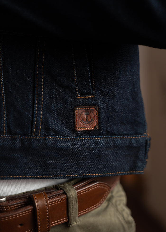 Close-up of a denim jacket with a leather patch above a brown belt and green pants, highlighting casual fashion details.