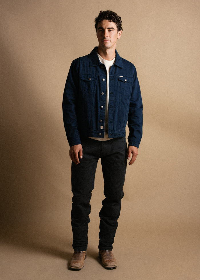 Man in a denim jacket and black jeans stands against a beige background, wearing brown boots, looking confidently ahead.