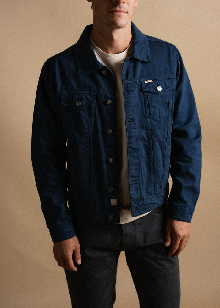 Man in a blue denim jacket and white shirt stands against a beige background, hands relaxed by his sides, exuding casual style.