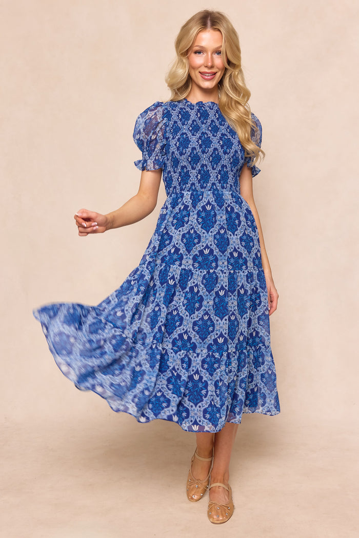 A woman with long blonde hair smiles and twirls in the Ivy City Co Cordelia Dress in Trellis Foulard—this blue patterned chiffon midi is perfect for a garden party—styled with beige heeled sandals against a neutral background.
