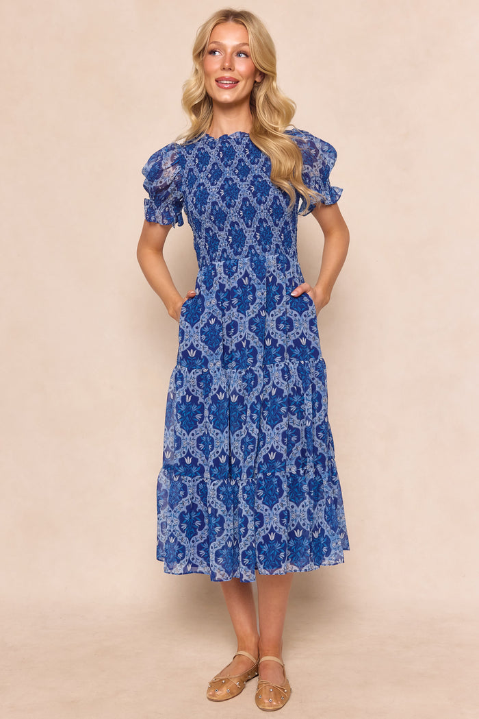 A woman smiles in the Ivy City Co Cordelia Dress in Trellis Foulard, a blue patterned chiffon midi with short puff sleeves, tiered skirt, and pockets. She has long blonde hair and tan flats against a beige background. Maternity friendly style.