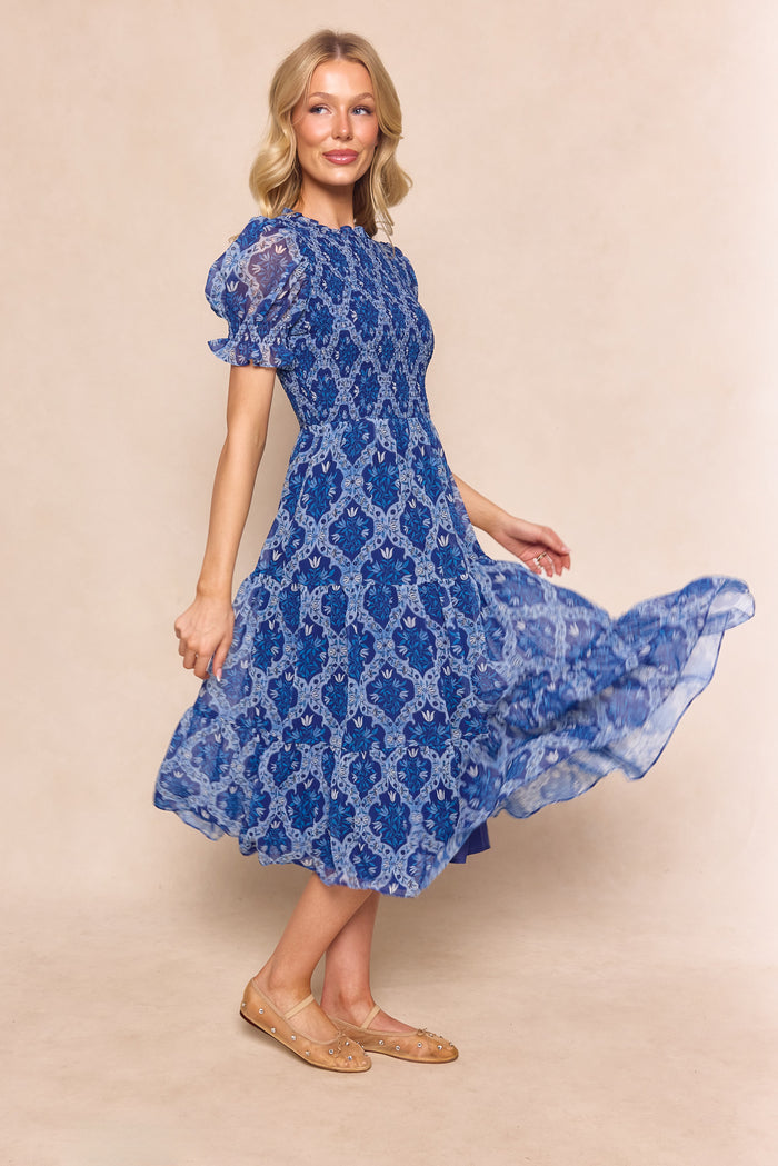 A woman in the Ivy City Co Cordelia Dress in Trellis Foulard, a blue patterned chiffon midi with a fitted waist and flowing skirt, stands smiling and twirling against a neutral background, styled with tan flats for an effortlessly chic look.