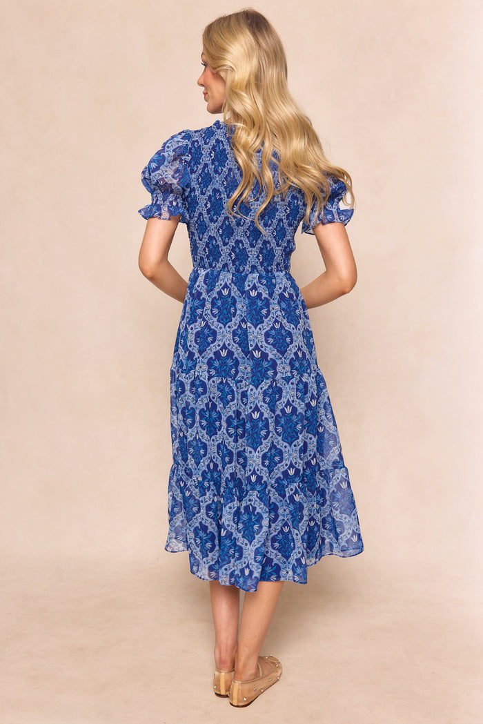 A woman with long blonde hair, facing away, wears the Ivy City Co Cordelia Dress in Trellis Foulard—a blue patterned chiffon maternity-friendly midi—paired with beige sandals, set against a light beige background.