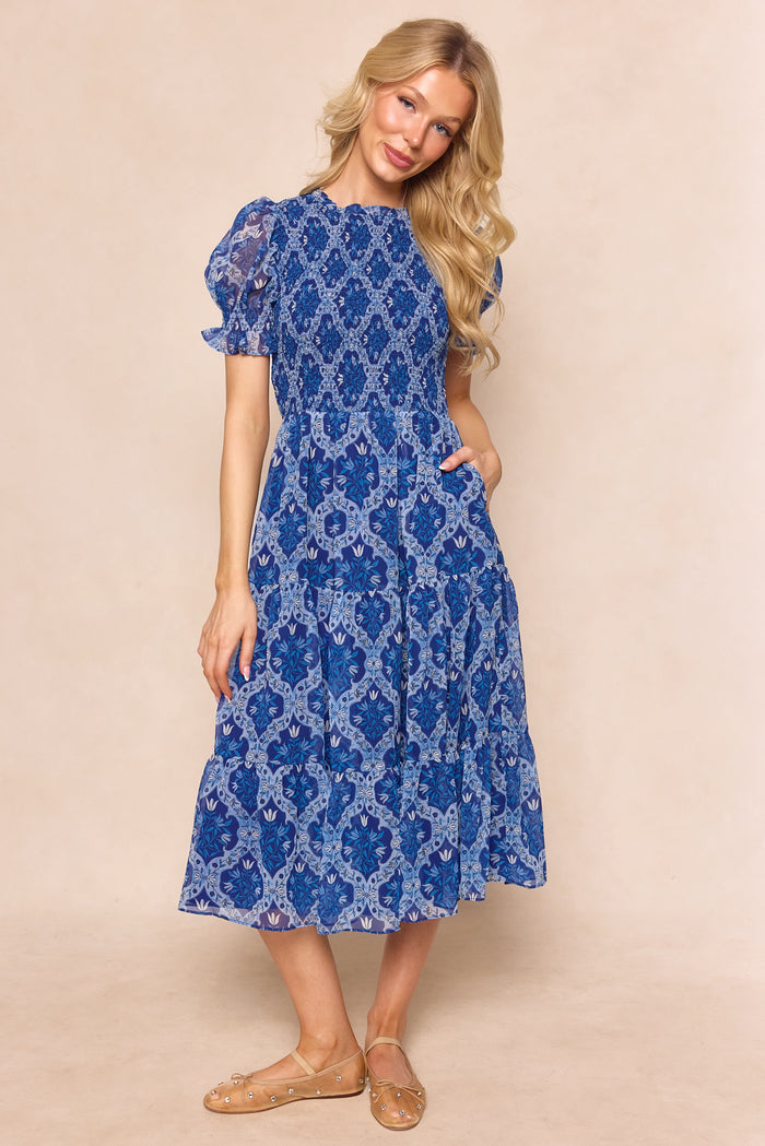 A woman with long blonde hair wears the Ivy City Co Cordelia Dress in Trellis Foulard, a blue chiffon maternity-friendly midi with an intricate white pattern, puff sleeves, and flat beige shoes—perfect for a garden party.