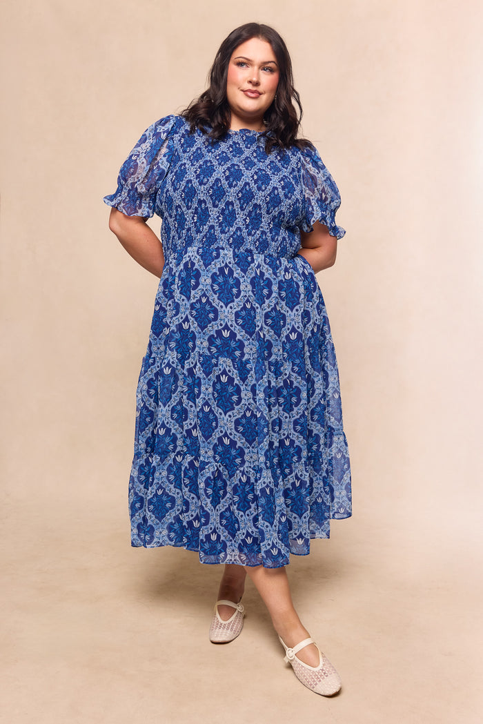 A woman with dark hair models the Ivy City Co Cordelia Dress in Trellis Foulard—a blue, patterned chiffon midi with short puff sleeves—paired with white woven shoes, making it a charming garden party choice against a neutral backdrop.