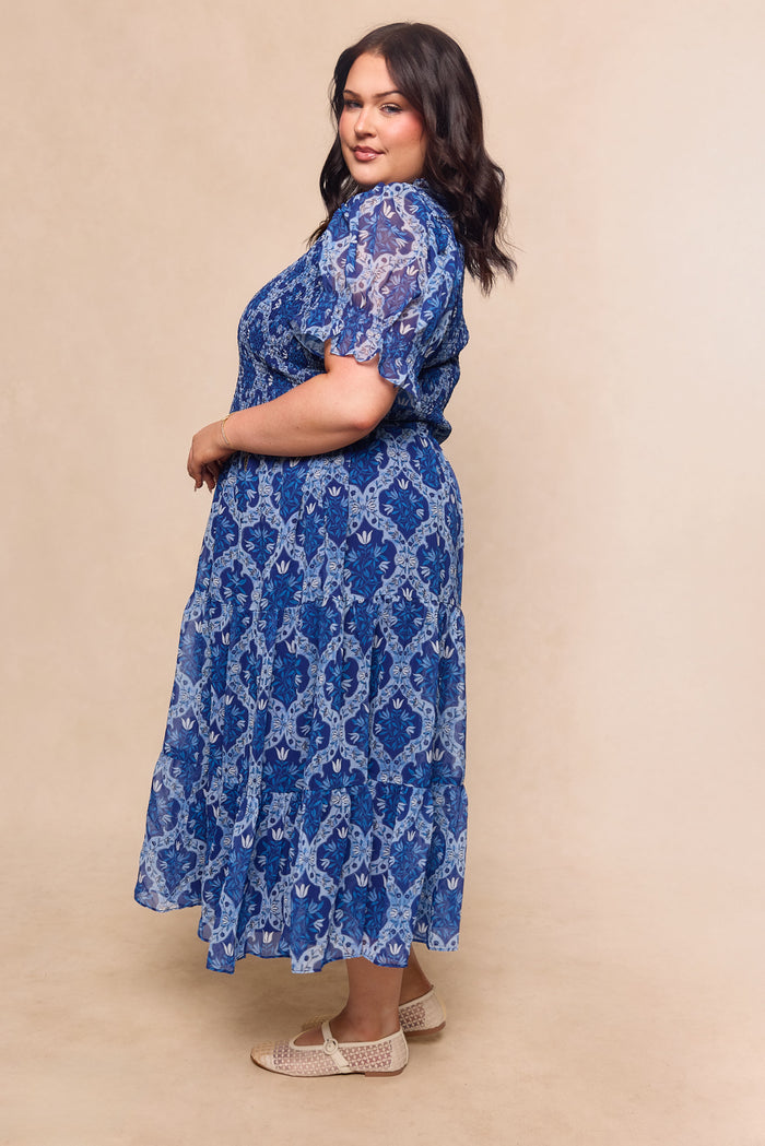 A woman with dark hair wears the Ivy City Co Cordelia Dress in Trellis Foulard—an elegant blue patterned chiffon midi—paired with beige woven shoes, standing sideways and smiling, making it a maternity-friendly garden party choice.
