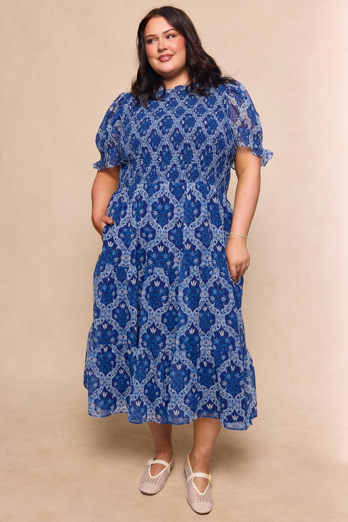 A woman in the Ivy City Co Cordelia Dress in Trellis Foulard, a blue patterned chiffon midi with short puff sleeves, stands smiling against a beige background. Styled maternity friendly with light shoes and dark hair.