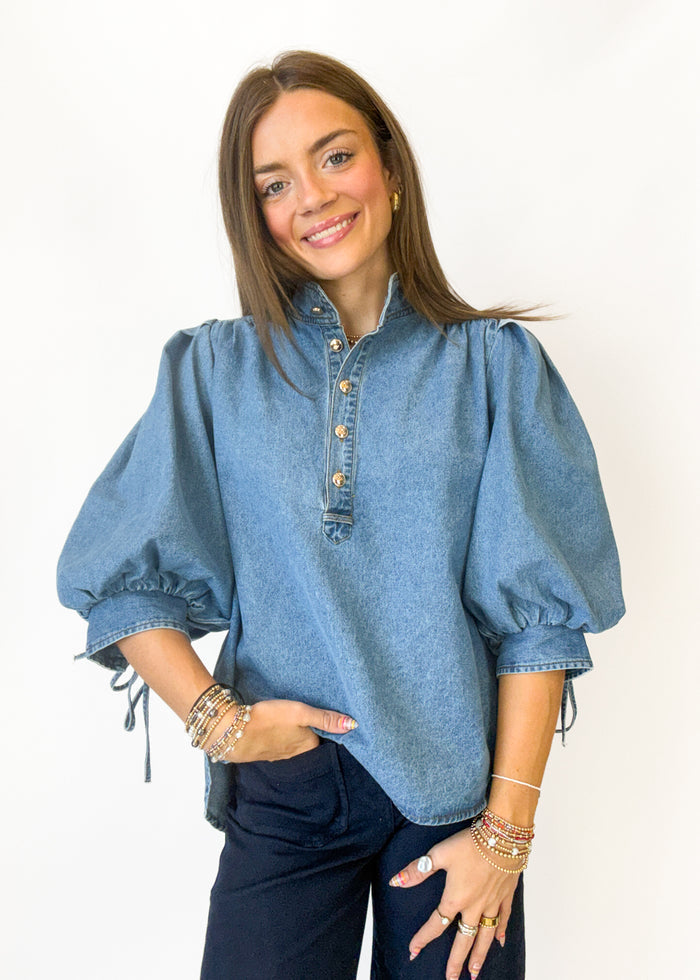 A woman modeling a light blue denim puff-sleeve blouse with a buttoned stand collar and dark navy pants, standing against a plain white background.