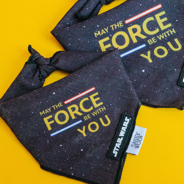 The Sassy Woof Dog Bandana - STAR WARS™ The Force features a black space pattern, "May the Force Be With You" in yellow, and Star Wars tags. Made of lightweight cotton, it fits necks up to 21.5" (sizes S-L), on a bright yellow background.