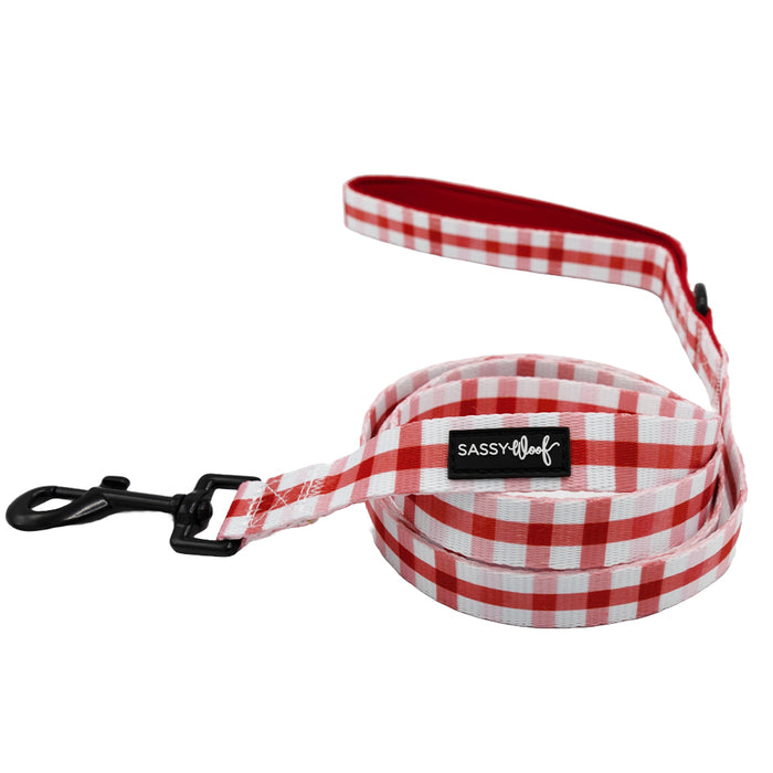 The Sassy Woof Dog Leash - Picnic Fur Two features a red and white checkered design, padded handle, black metal clip, and logo tag—coiled with the loop visible. Perfect to pair with a matching harness.