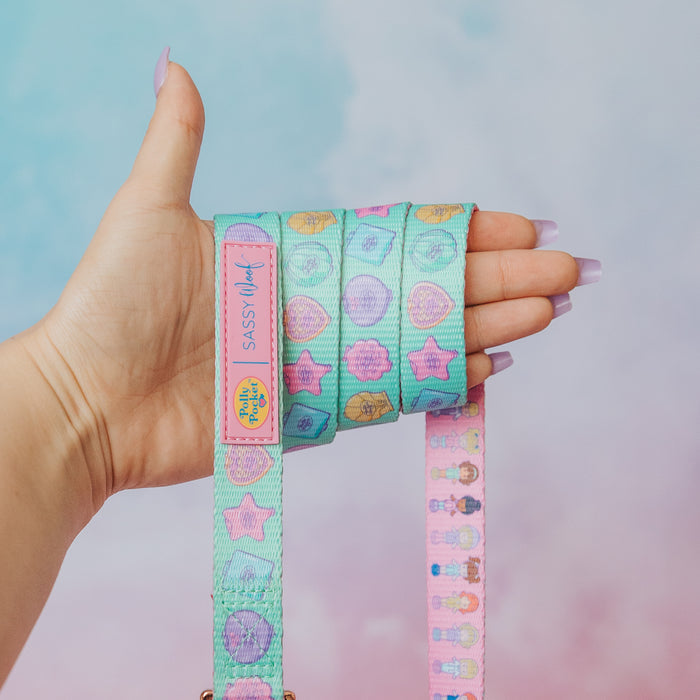 A hand with pastel purple nails holds the Sassy Woof Collar Three Piece Bundle - Polly Pocket™, decorated with cartoon stars, hearts, and circles. The colorful design pops against the pink and blue background.