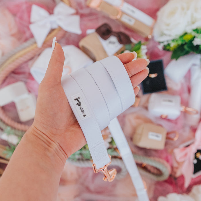 A hand holds the Sassy Woof Pup of Honor Dog Leash with a rose gold clasp and neoprene handle, surrounded by matching accessories, flowers, and pastel items on a pink background.