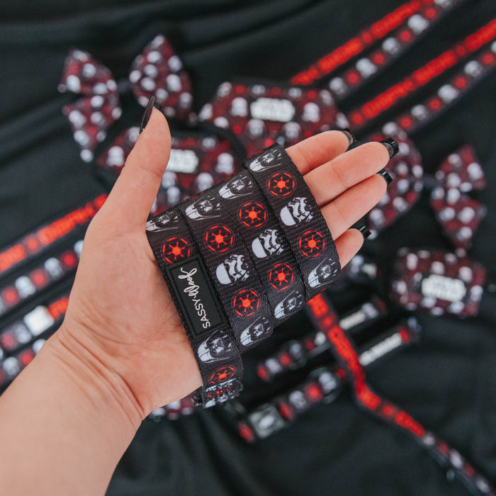 A hand holds the Sassy Woof Dog Leash - STAR WARS™ The Dark Side, featuring black material with red and white Empire logos and Stormtrooper helmets; matching bows and ribbons are blurred in the background.