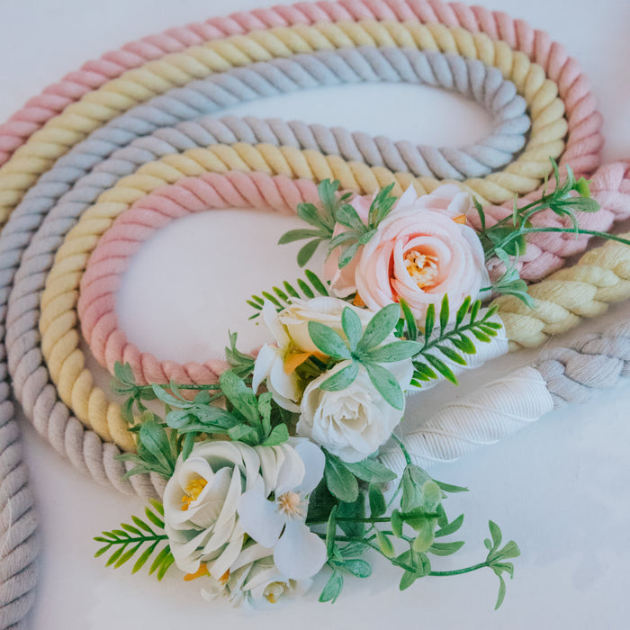 Three thick, twisted Sassy Woof dog leashes in pastel pink, yellow, and gray are looped on a white surface. Each leash features the Dog Leash Flower Accessory with artificial white and peach blooms plus green leaves for decoration.