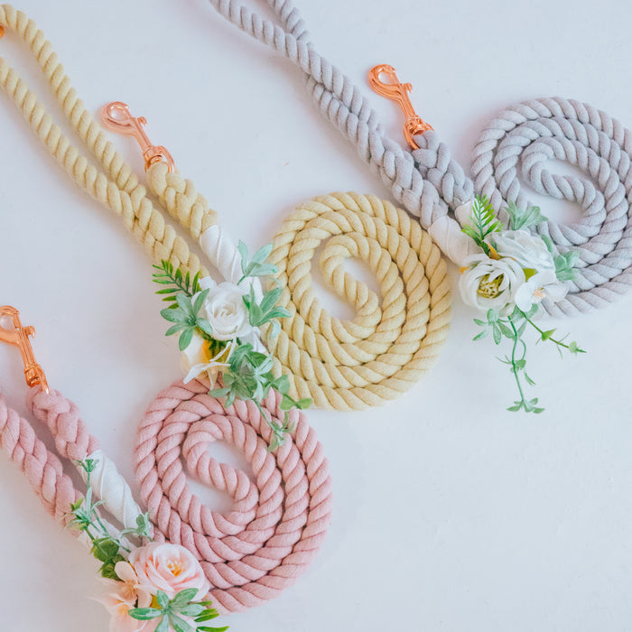 Three Sassy Woof coiled dog leashes in pastel pink, yellow, and gray, each adorned with a Dog Leash Flower Accessory of artificial white flowers and green leaves, are displayed on a white surface with rose gold metal clips.