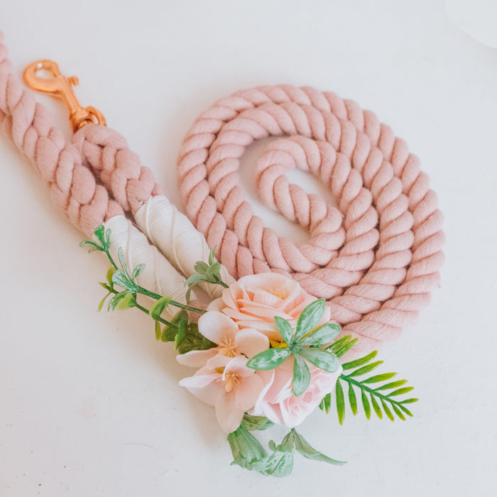 The Sassy Woof Dog Leash Flower Accessory is a coiled pale pink leash with a gold clasp, featuring artificial light pink flowers and green leaves at one end, displayed on a white surface.