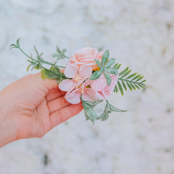 A hand holds a small bouquet of light pink flowers and green leaves, featuring the Sassy Woof Dog Leash Flower Accessory, set against a soft, blurred white background.