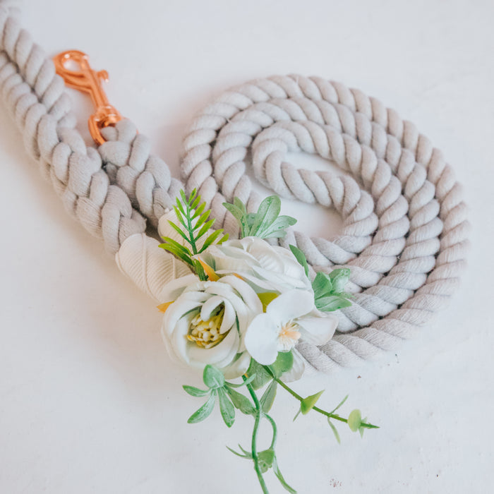 The Sassy Woof Dog Leash Flower Accessory, featuring artificial white blooms and green leaves, is displayed on a braided light gray leash with a rose gold clasp, coiled neatly on a white surface.