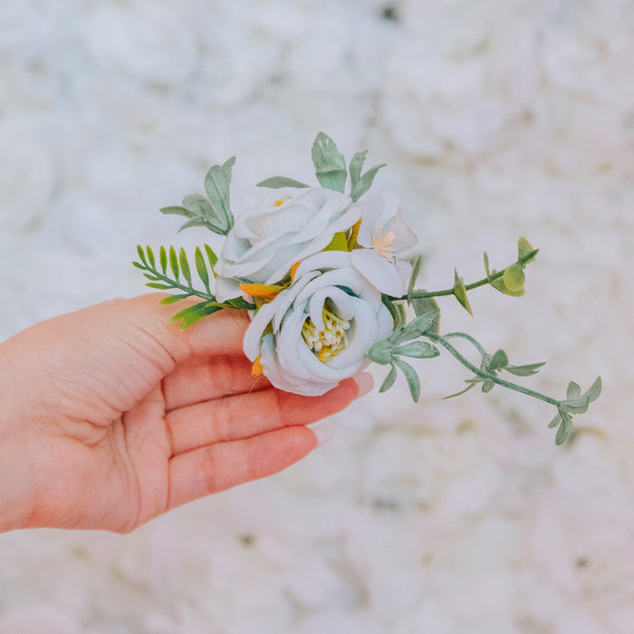 A hand holds Sassy Woof’s Dog Leash Flower Accessory, a small white rose and green leaf arrangement with a Velcro attachment, set against a soft, blurred floral background.