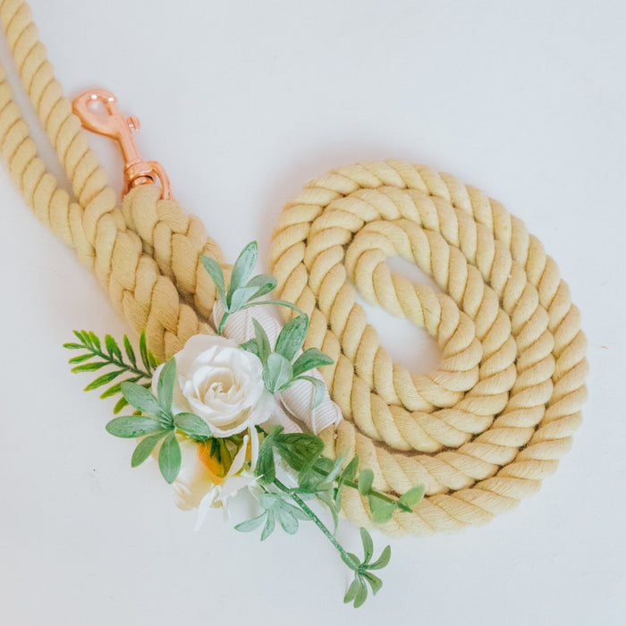 The Sassy Woof Dog Leash Flower Accessory features a coiled yellow leash with a rose gold clasp and artificial white flower blooms with green leaves, shown on a white surface.