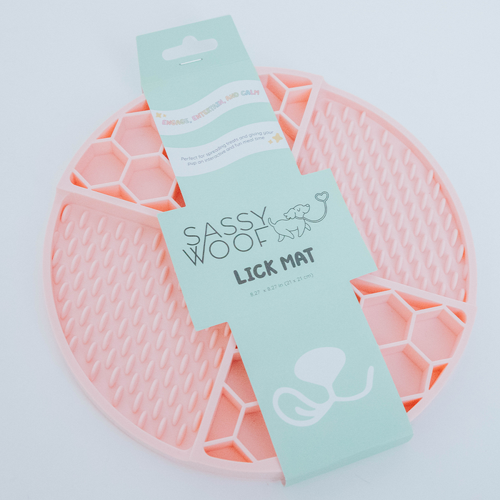 The Sassy Woof Dog Lick Mat is a round, pink silicone mat with honeycomb and dot patterns, shown in its packaging. It's designed to calm and entertain pets, making it ideal for relieving dog anxiety.
