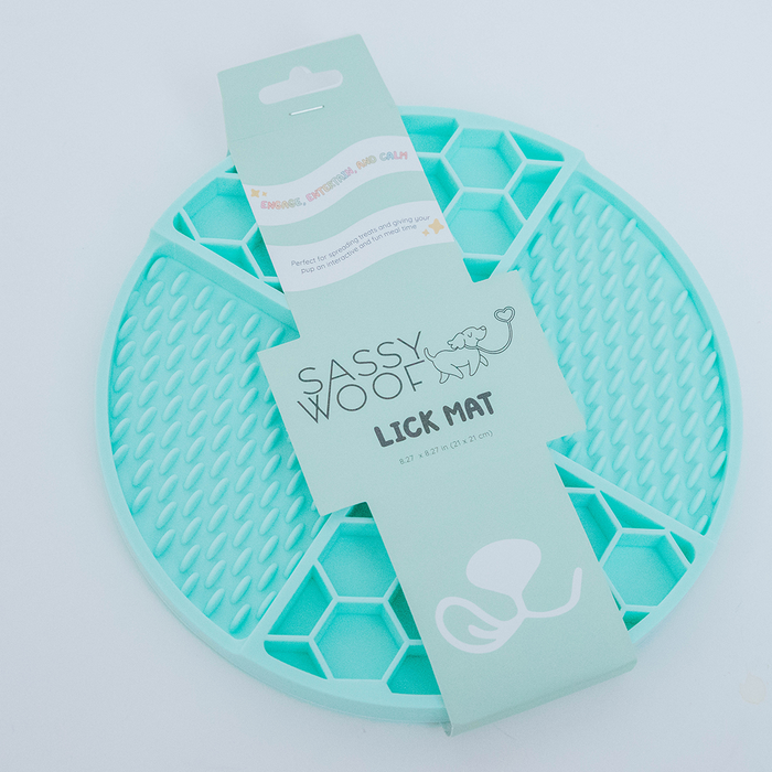 The Sassy Woof Dog Lick Mat is a round, teal silicone mat for dogs with honeycomb and dot textures. Packaged with a paper band, it features the brand name, a dog illustration, and an octopus graphic.