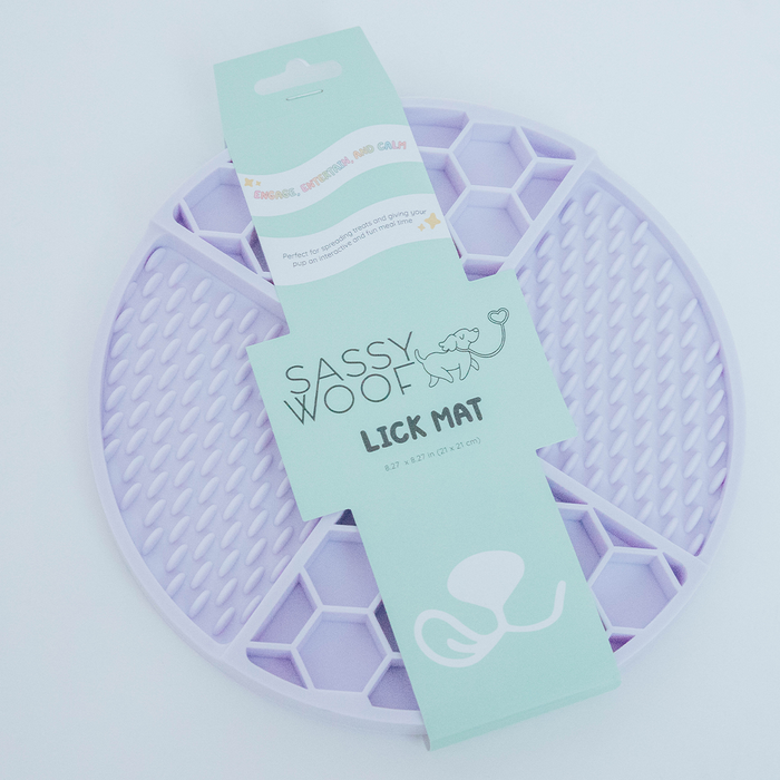 The Sassy Woof Dog Lick Mat is a round, light purple silicone mat with a honeycomb texture, shown in packaging with pastel green labels—a fun interactive toy designed to help relieve dog anxiety.