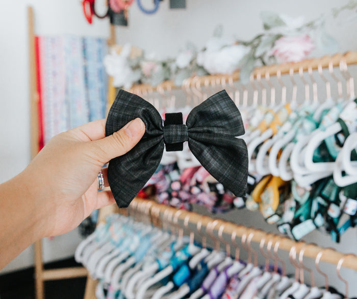 A hand holds the Sassy Woof Dog Sailor Bow - Baby Got Black in front of a boutique rack filled with colorful bows, dog bows, and headbands hanging on display.