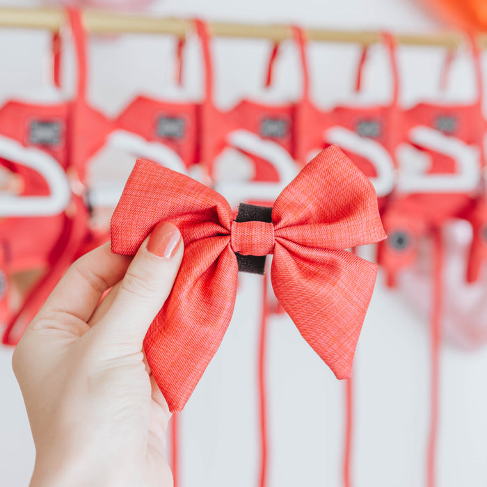 A hand with painted nails holds the Sassy Woof Dog Sailor Bow - Merlot in focus, while several matching red pieces from the Merlot collection hang blurred in the background.