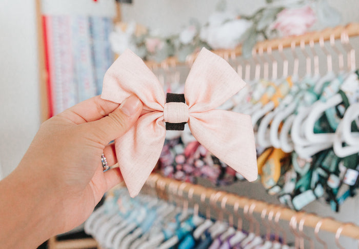 A hand holds the Sassy Woof Dog Sailor Bow - Pinot in light pink with a black center, in front of a rack showcasing colorful, patterned bows and accessories from the Pinot collection.