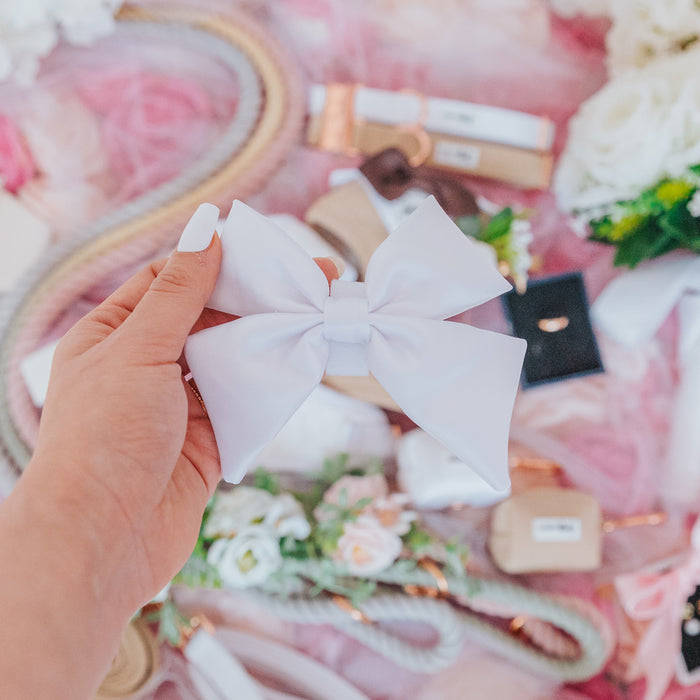 A hand with white-painted nails holds the "Dog Sailor Bow - Pup of Honor" by Sassy Woof. Surrounded by matching dog accessories and pastel flowers, it's perfect for a coordinated dog wedding outfit.
