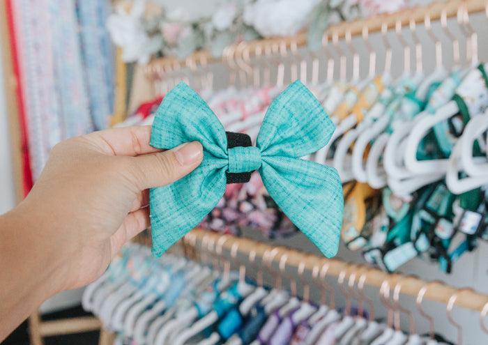A hand holds the Sassy Woof Dog Sailor Bow - Wag Your Teal, featuring a turquoise bow with a black band, in front of racks displaying colorful dog bows and accessories on hangers.