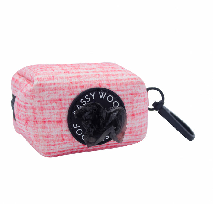 The Sassy Woof Dog Waste Bag Holder - Dolce Rose features a pink and white checkered design, black circular opening, partially pulled-out bag, and a black clip for attaching to your leash or dog accessories.
